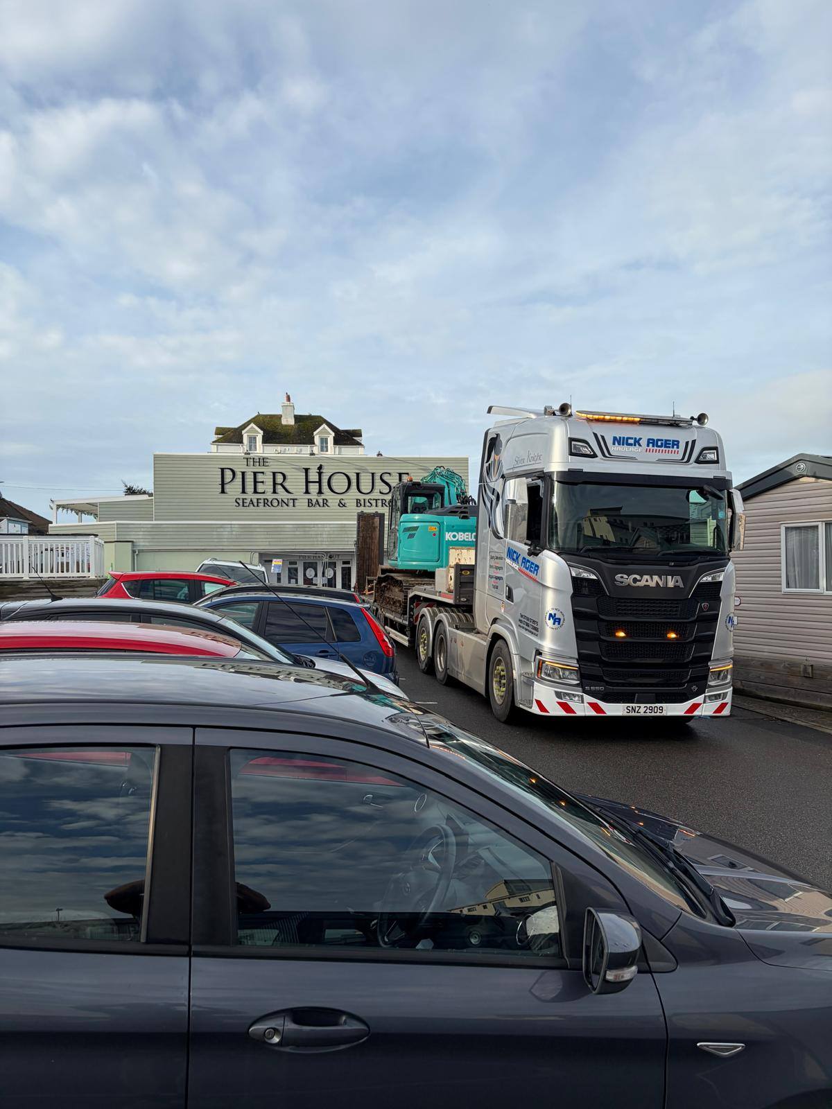Scania Truck in Front of The Pier House Seafront Bar A Scania truck parked in front of The Pier House seafront bar and bistro, with several cars in the foreground and a blue piece of machinery visible behind the truck.