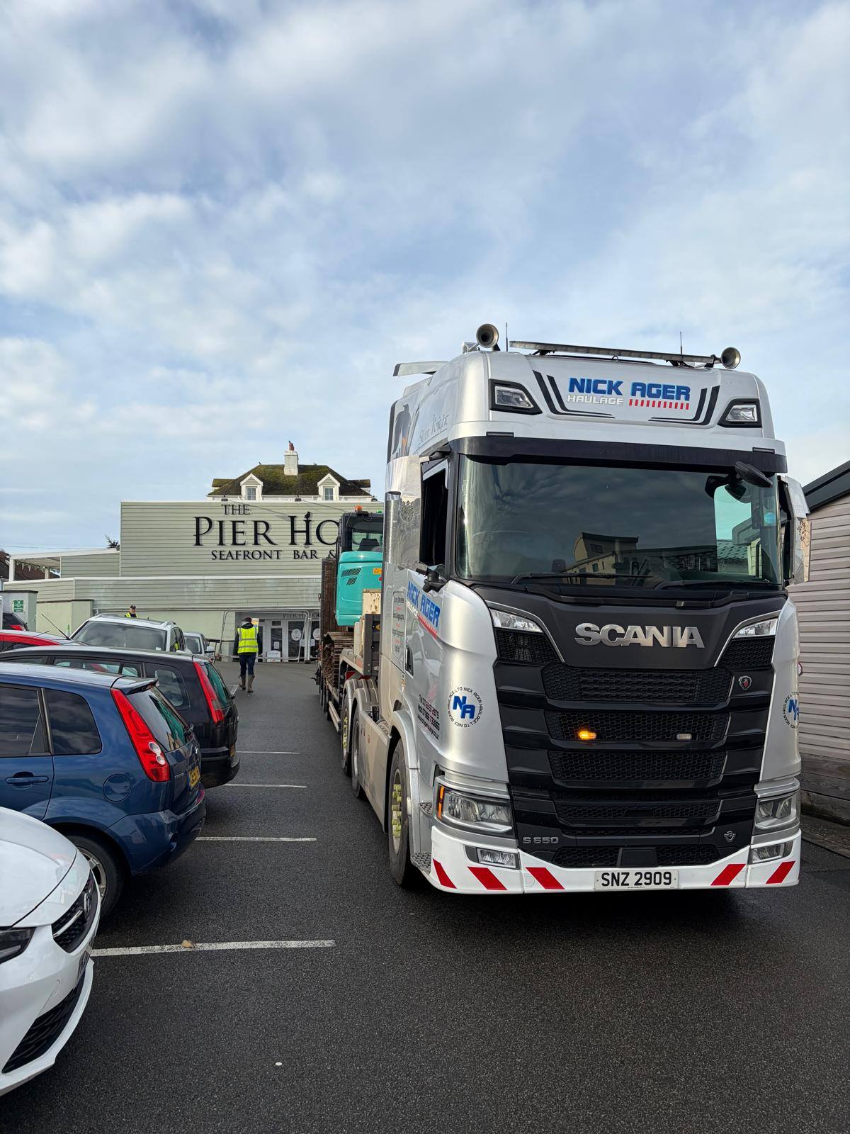 A Scania truck parked in front of The Pier House seafront bar and bistro, with several cars in the foreground and a blue piece of machinery visible behind the truck.