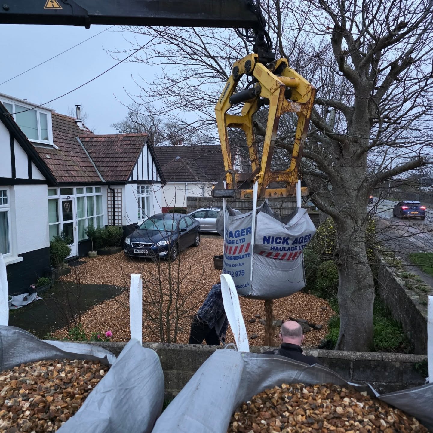 Heavy Machinery Delivering Aggregate to Residential Area A yellow crane lifting heavy bags of aggregate while parked cars and a residential house are visible in the background.