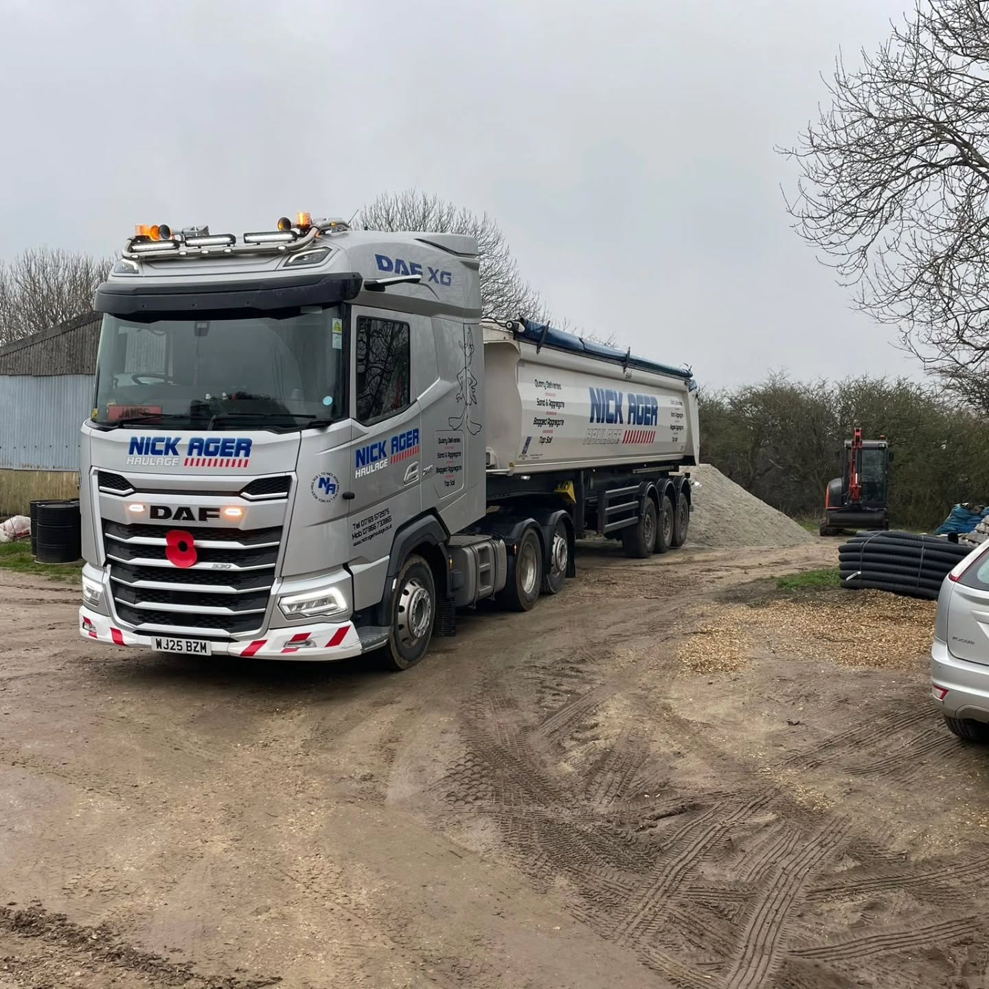 DAF XG Truck in an Industrial Setting A DAF XG truck belonging to Nick Ager Haulage is parked on a gravel surface with a trailer loaded with raw materials. In the background, a small excavator and storage items are visible.