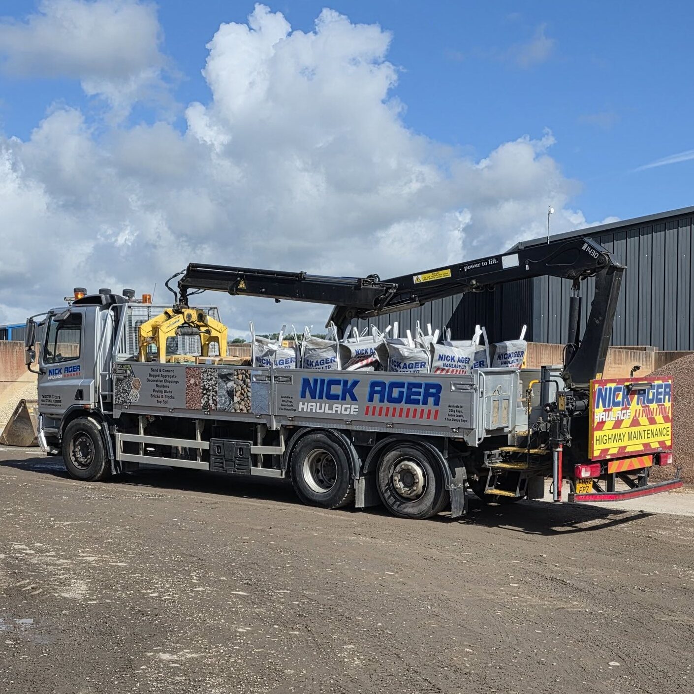 NAH Dumpy Bags A Nick Ager Haulage truck equipped with a crane, loaded with bags of materials, parked at a construction site under a blue sky with fluffy clouds.