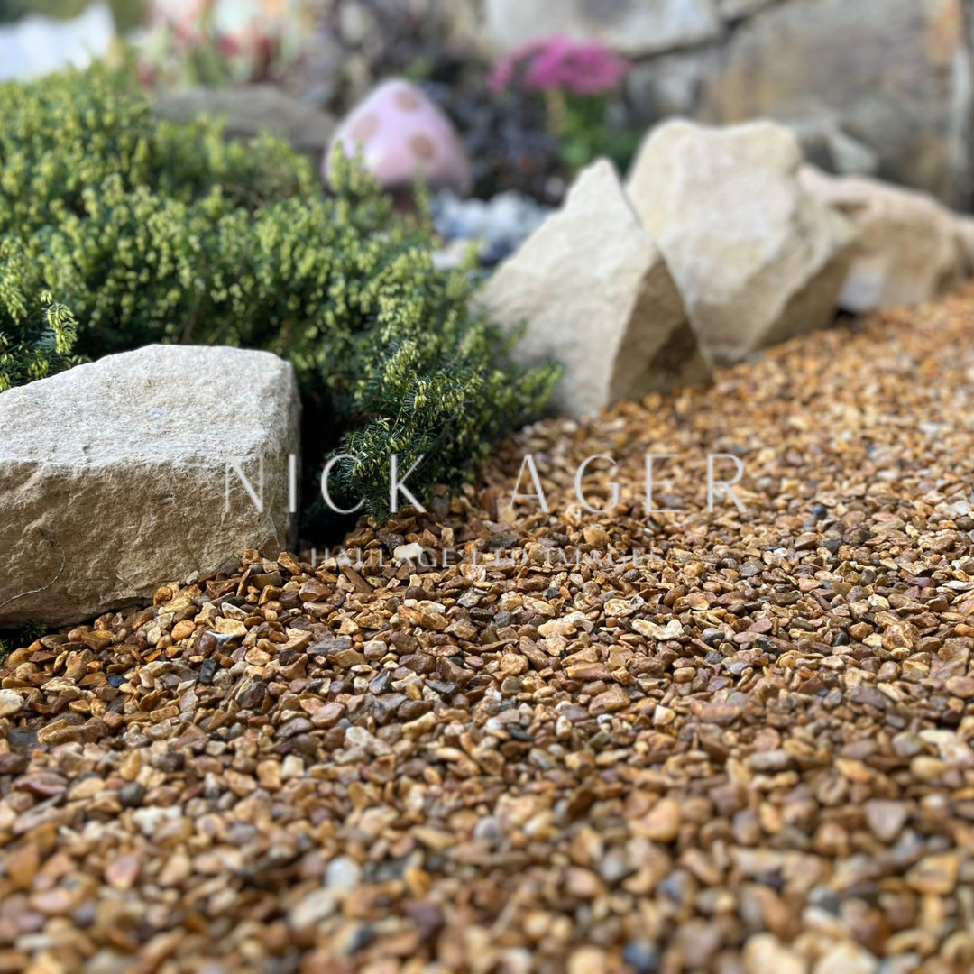 Close-up of gravel landscaping featuring various sizes of brown pebbles, bordered by large natural stones and lush green foliage.