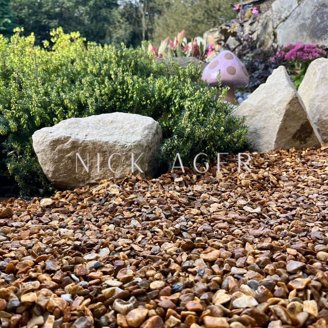 A view of a garden featuring a gravel pathway made of small stones, surrounded by lush greenery and decorative stones, with a whimsical pink mushroom peeking in the background.