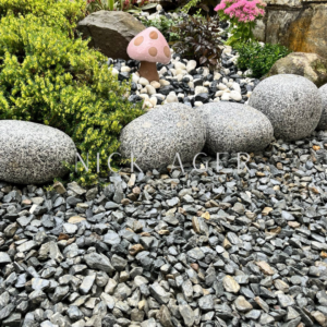 A serene garden scene featuring smooth grey stones arranged along gravel, vibrant green plants, and a whimsical pink mushroom decoration in the background.