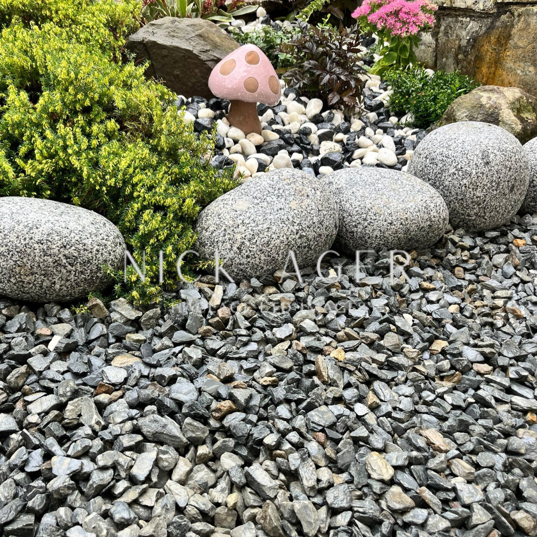 A serene garden scene featuring smooth grey stones arranged along gravel, vibrant green plants, and a whimsical pink mushroom decoration in the background.