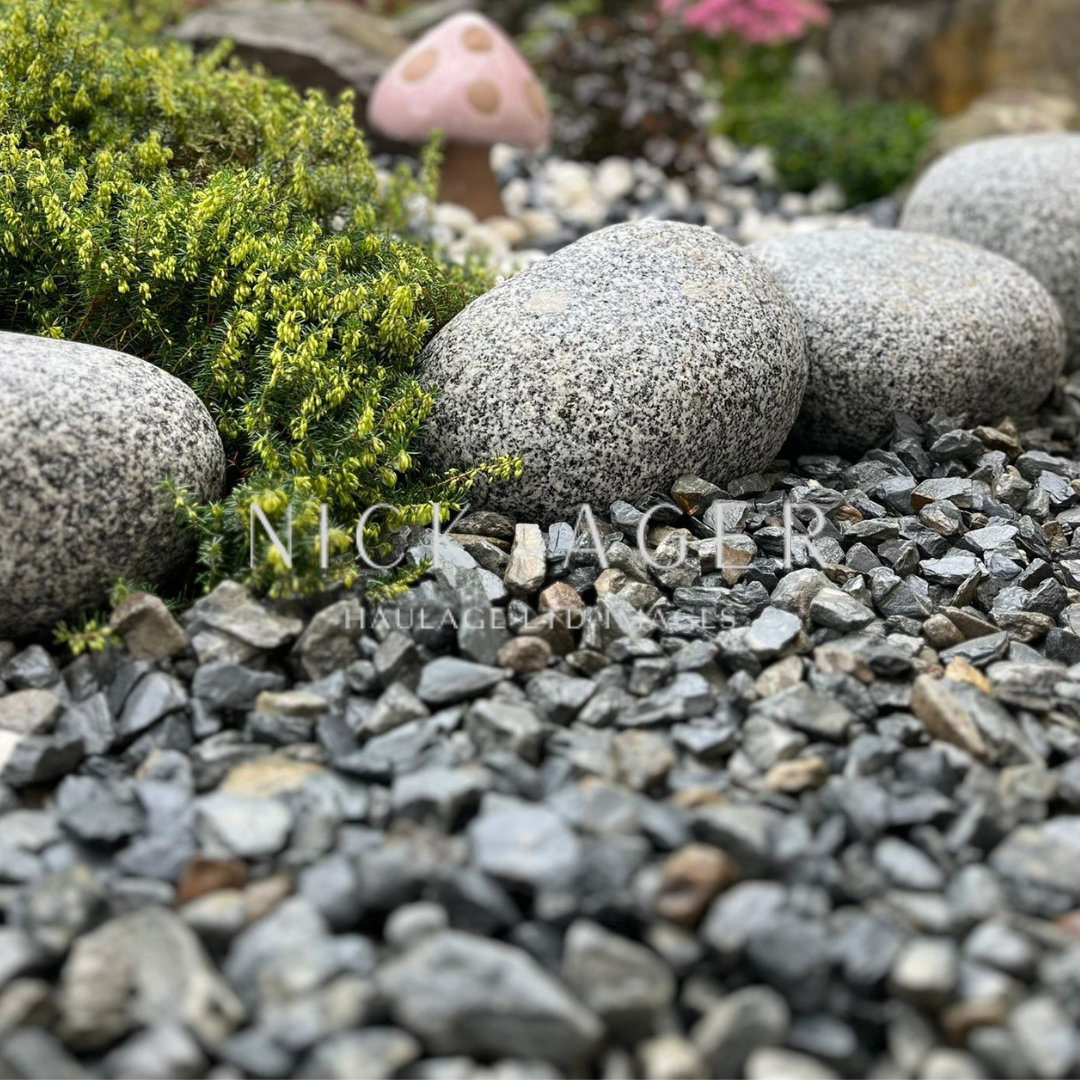 A close-up view of a garden landscape featuring smooth grey stones, small gravel, and lush green foliage, with a whimsical pink mushroom in the background.