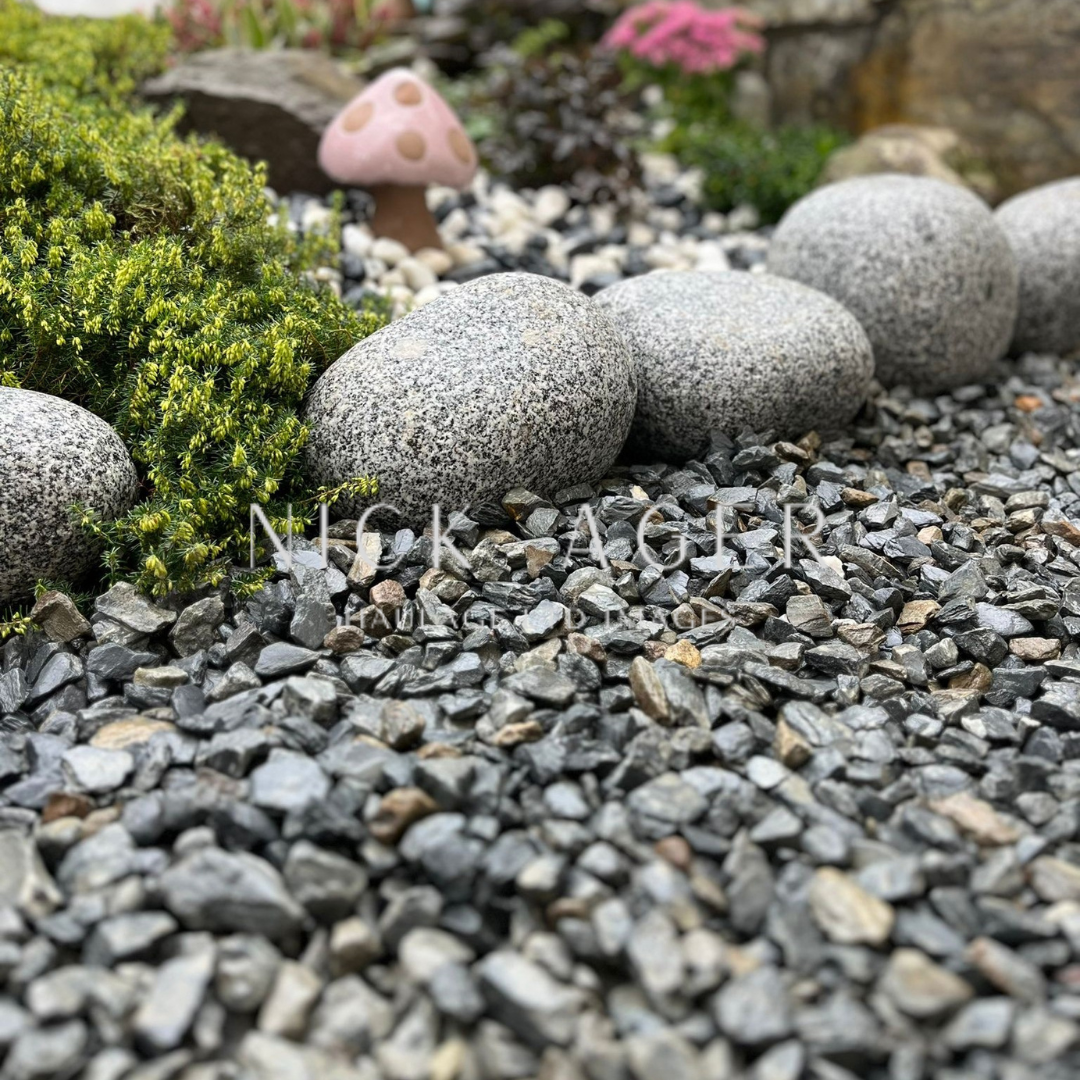 A garden view featuring smooth grey stones, decorative gravel, lush green foliage, and a pink mushroom ornament.