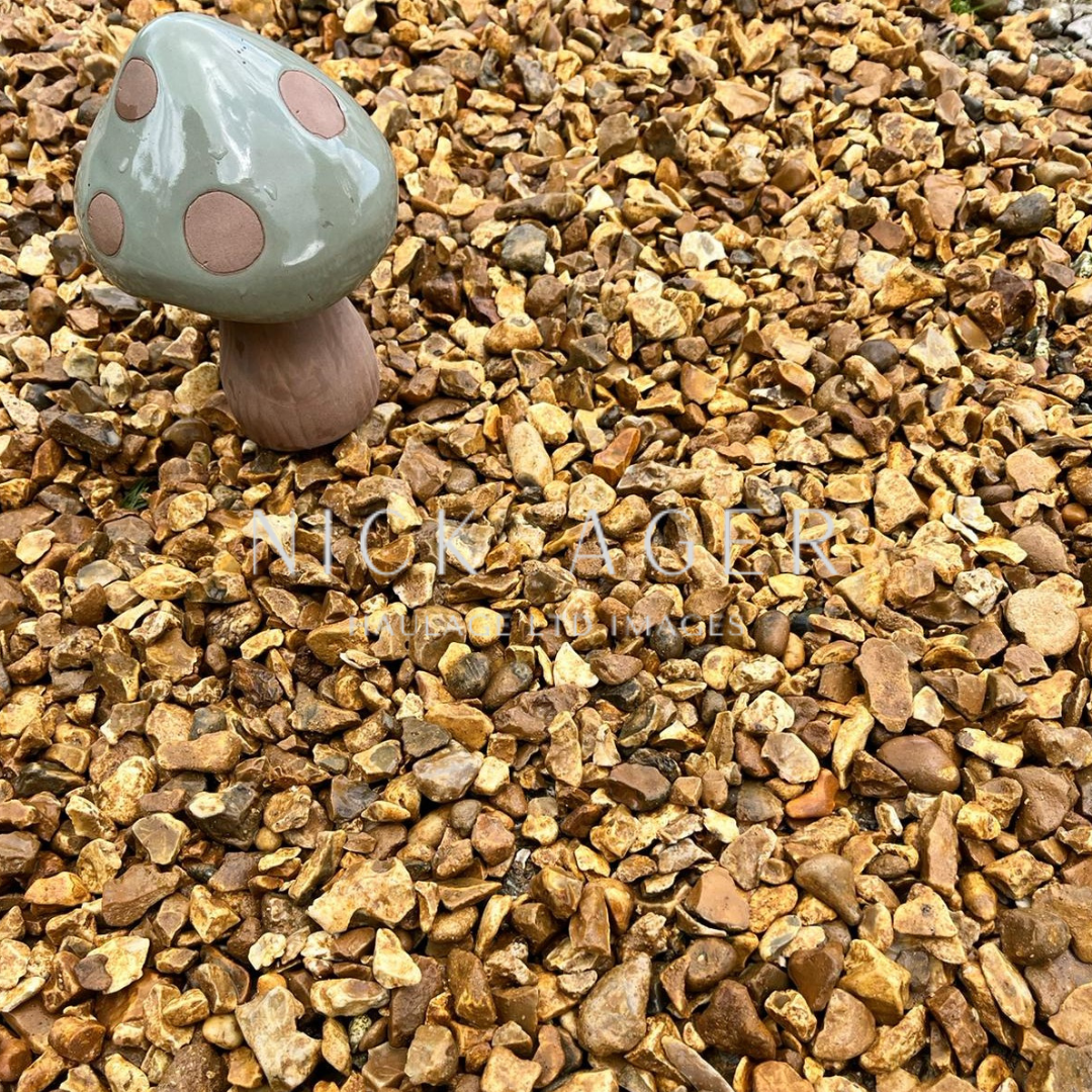 A decorative ceramic mushroom with a grey top and brown spots stands on a bed of golden gravel stones.