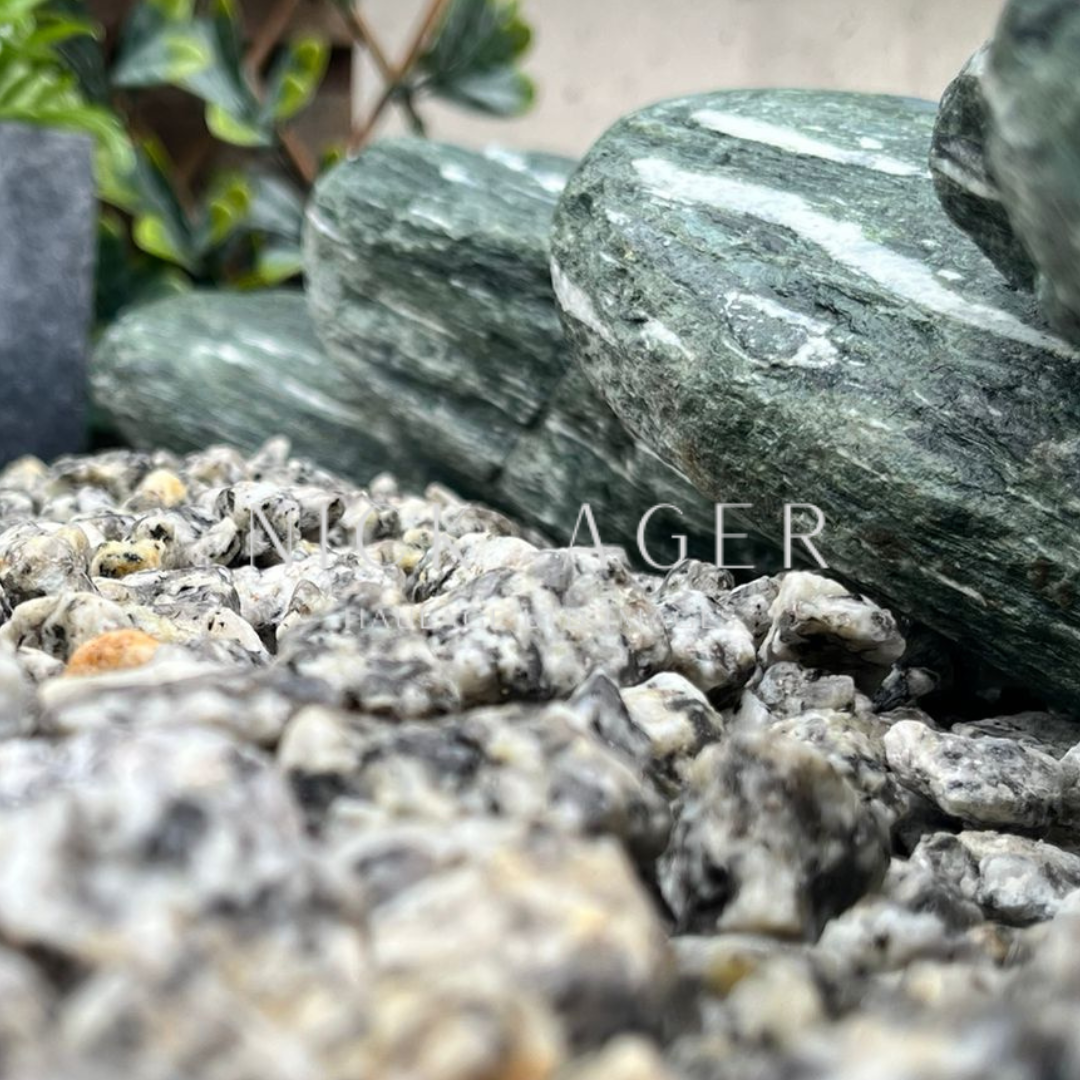 A close-up view of smooth green stones on top of grey and white gravel, with a blurred background of greenery.