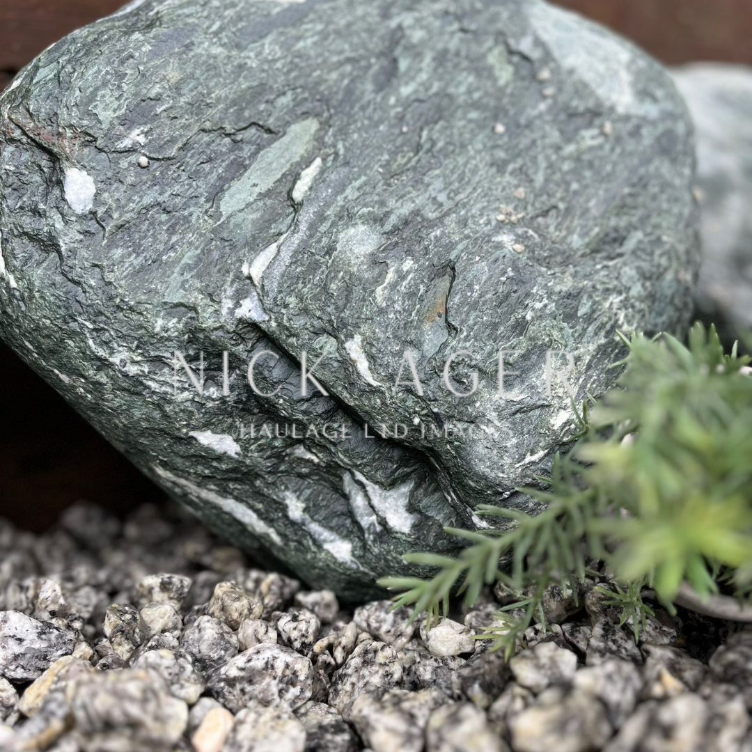 A close-up view of a large green stone with white streaks, surrounded by small gravel pebbles and green foliage.