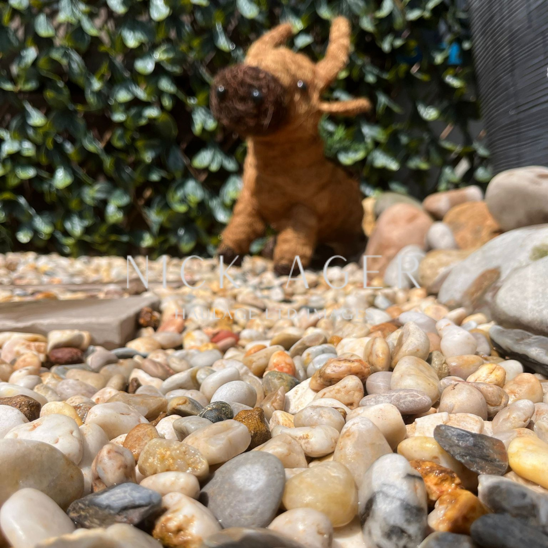 A woven animal figurine, resembling a moose, positioned among colourful pebbles in a garden, with green foliage in the background.