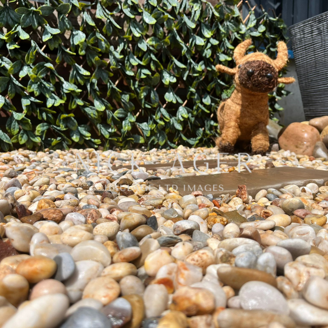 A felted animal resembling a small cow stands on a bed of smooth pebbles, with a backdrop of lush green foliage.