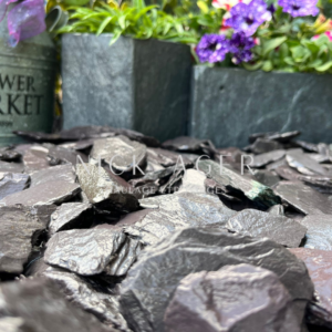 Close-up view of dark slate stones on the ground, with flower pots featuring green plants and purple flowers in the background.