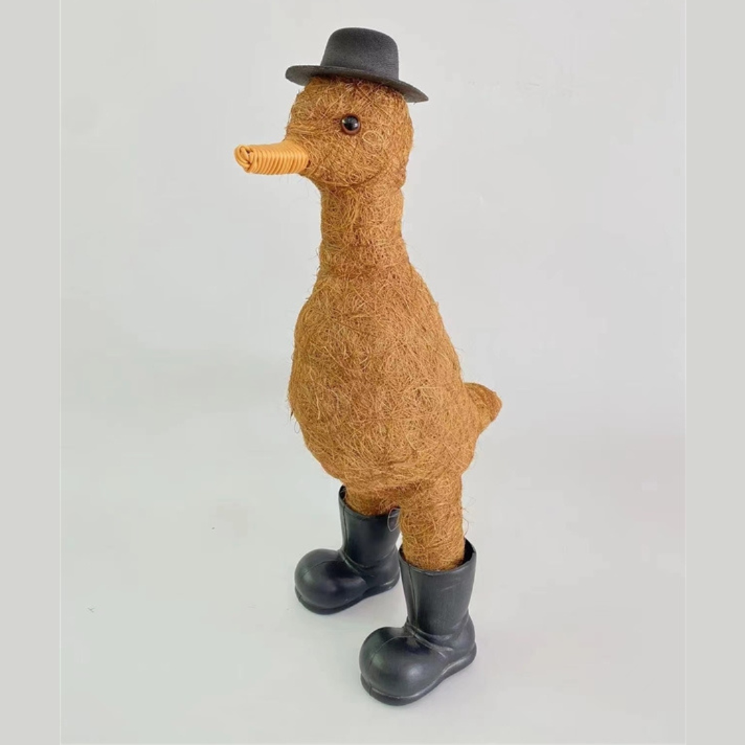 A whimsical decorative duck sculpture made from straw-like material, wearing a black hat and rubber boots, standing against a plain background.