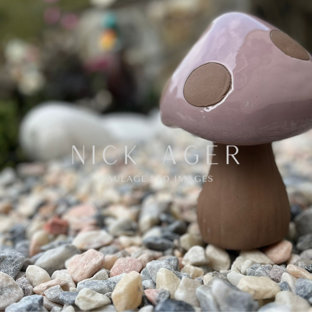 A close-up view of a decorative mushroom ornament with a pink cap and brown spots, sitting on a bed of small stones in a garden setting.
