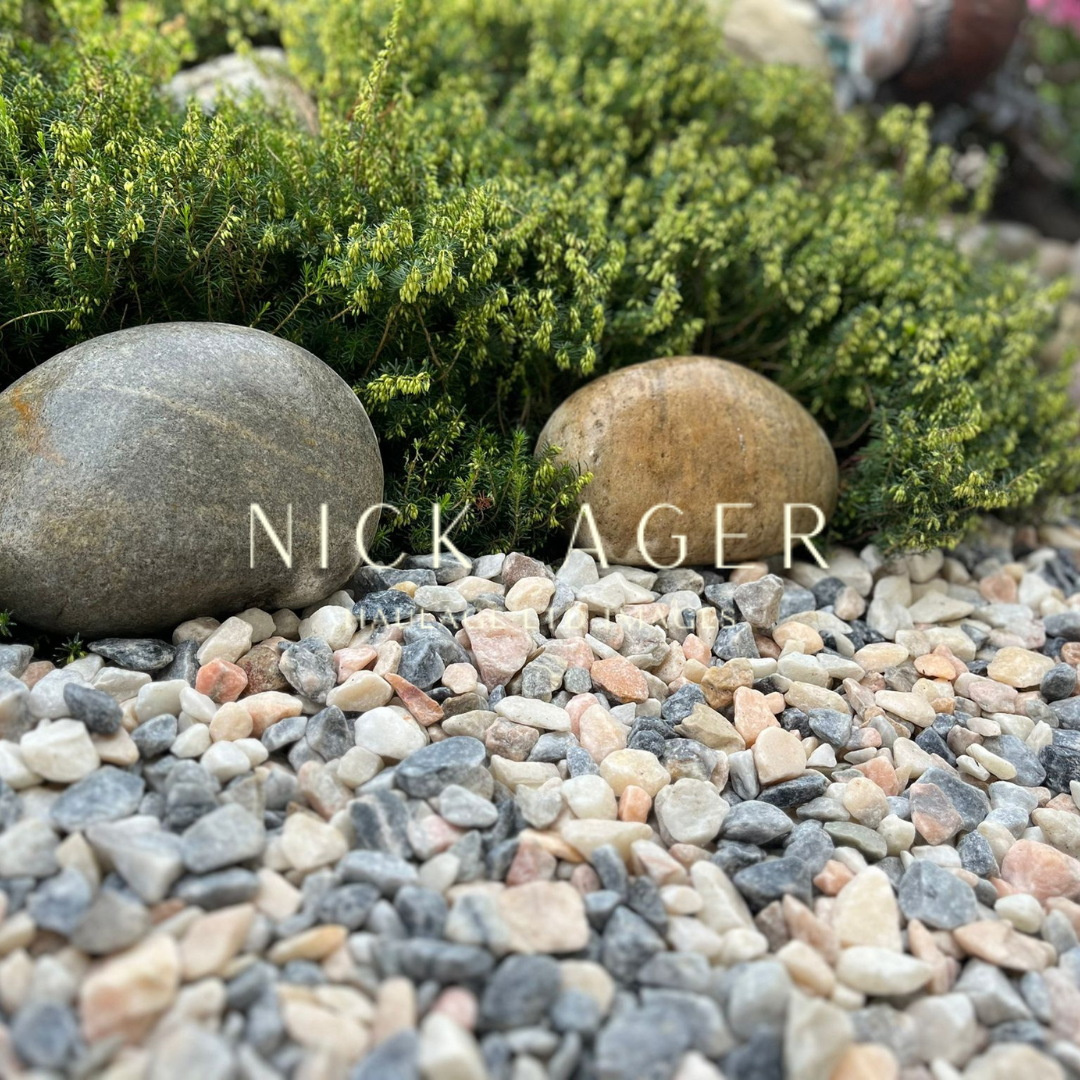 A close-up of a garden scene featuring smooth grey and brown rocks surrounded by small pebbles in shades of grey, pink, and white, with lush green plants in the background.