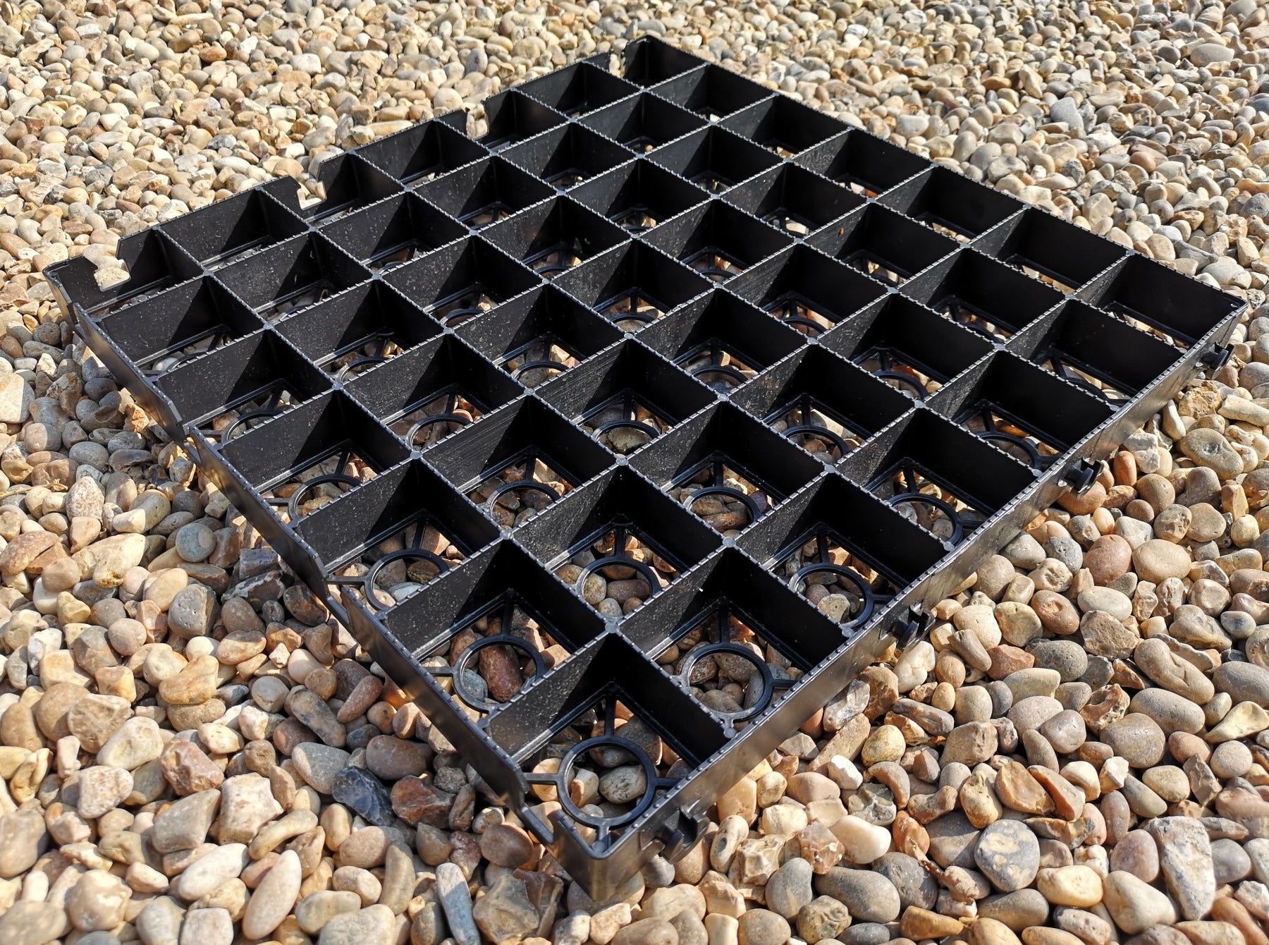 A black plastic planting tray designed with multiple square compartments, resting on a bed of small gravel stones.