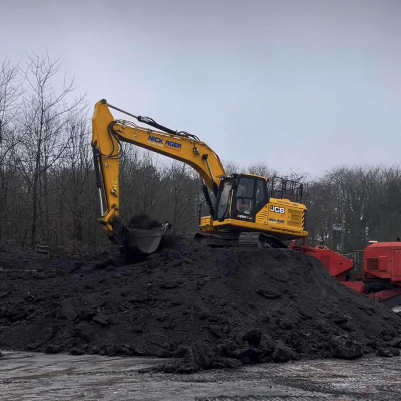 Excavator Operating on Construction Site A yellow JCB excavator is digging into a large mound of dark material on a construction site, with trees in the background and a muddy ground.