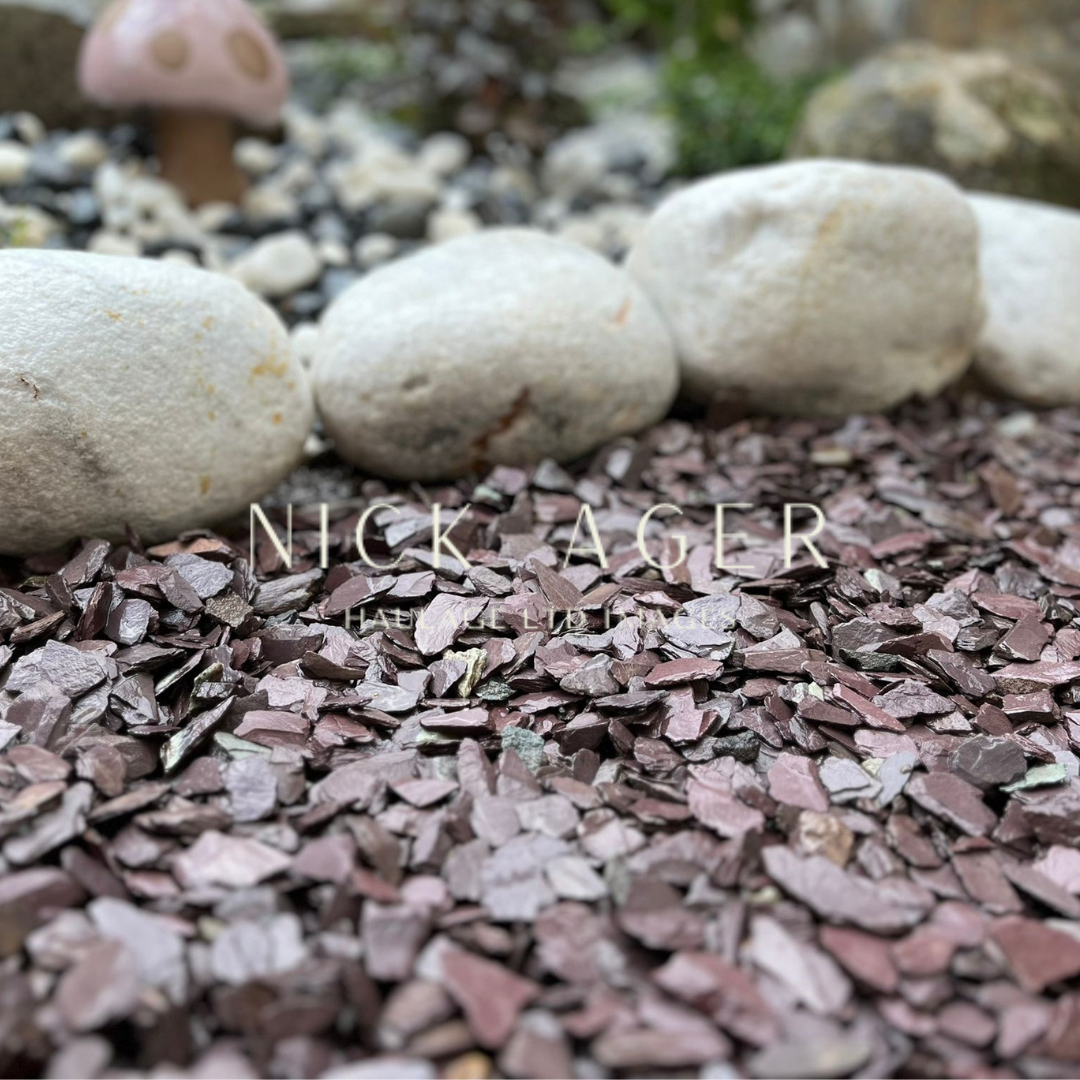 A close-up view of a garden area featuring decorative purple slate stones, contrasted by smooth white pebbles and a blurred background of greenery, including a small mushroom ornament.