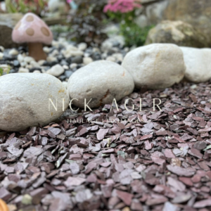 A close-up view of decorative garden landscaping featuring smooth white stones and maroon slate mulch, with a whimsical pink mushroom in the background.
