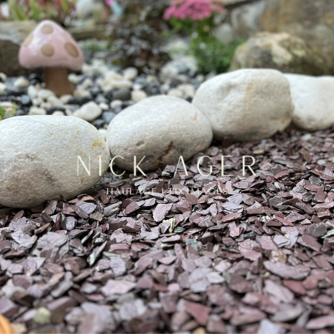 A close-up view of decorative garden landscaping featuring smooth white stones and maroon slate mulch, with a whimsical pink mushroom in the background.