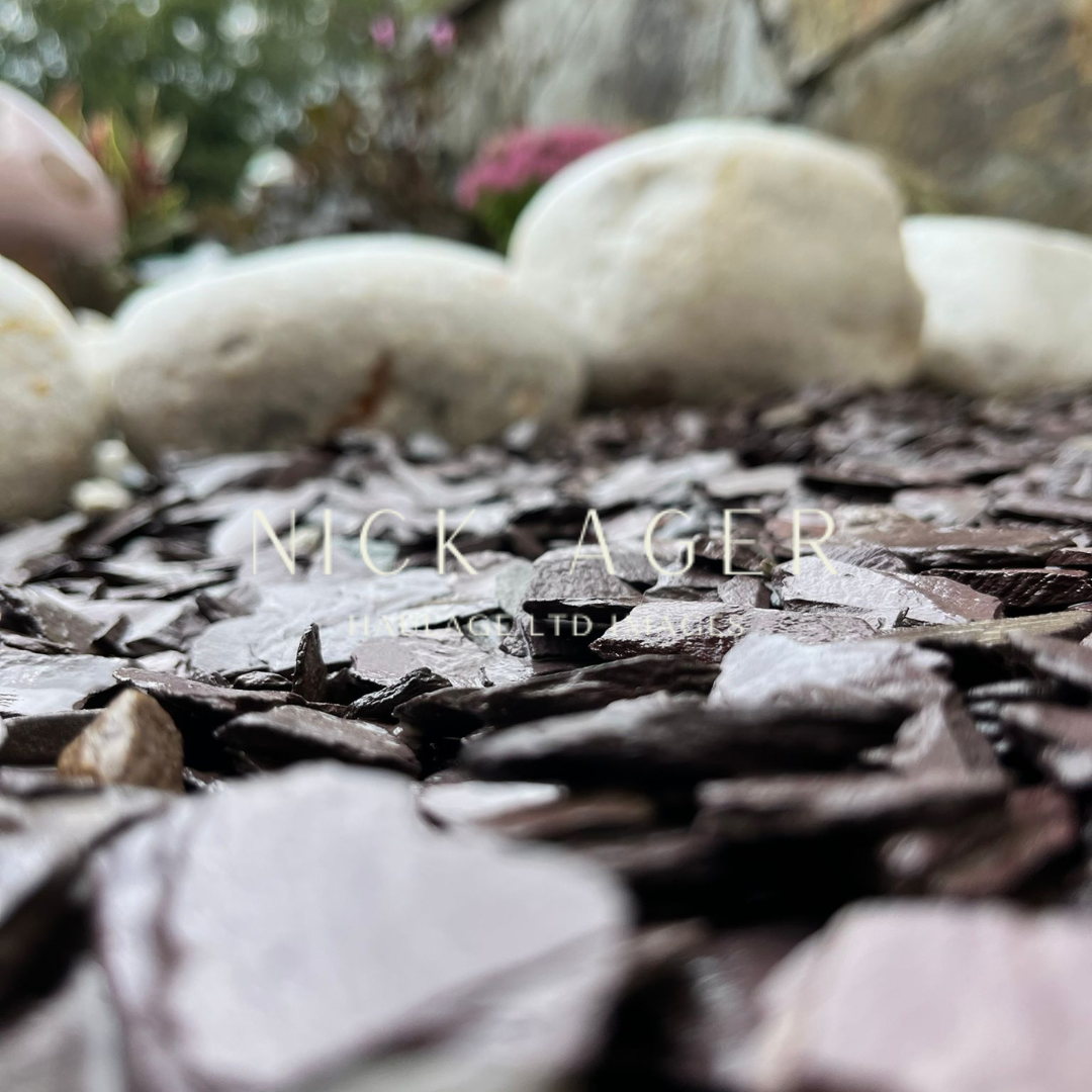 A close-up view of dark slate chips on the ground, surrounded by white decorative stones, with a blurred background of plants and flowers.