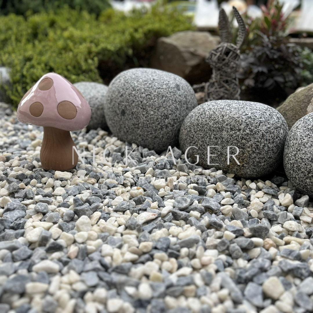 A decorative pink and brown mushroom figurine surrounded by grey and white pebbles, with larger grey stones in the background and a woven rabbit figure partially visible.