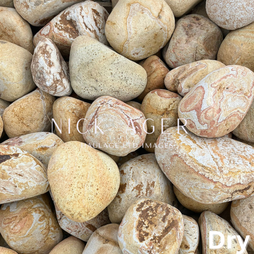 A close-up view of smooth, rounded pebbles featuring various shades of beige, brown, and cream with streaks of different colours and patterns, stacked together on a dry surface.