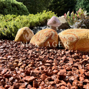 A close-up view of a garden featuring smooth decorative stones, a pink mushroom sculpture, and lush greenery. The ground is covered with reddish gravel.