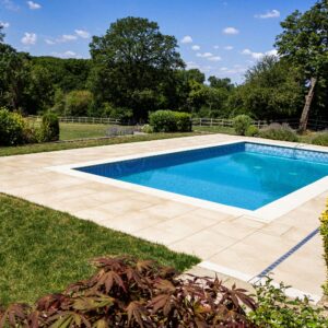 A sparkling blue swimming pool surrounded by a stone patio and vibrant greenery, under a clear blue sky with fluffy clouds.