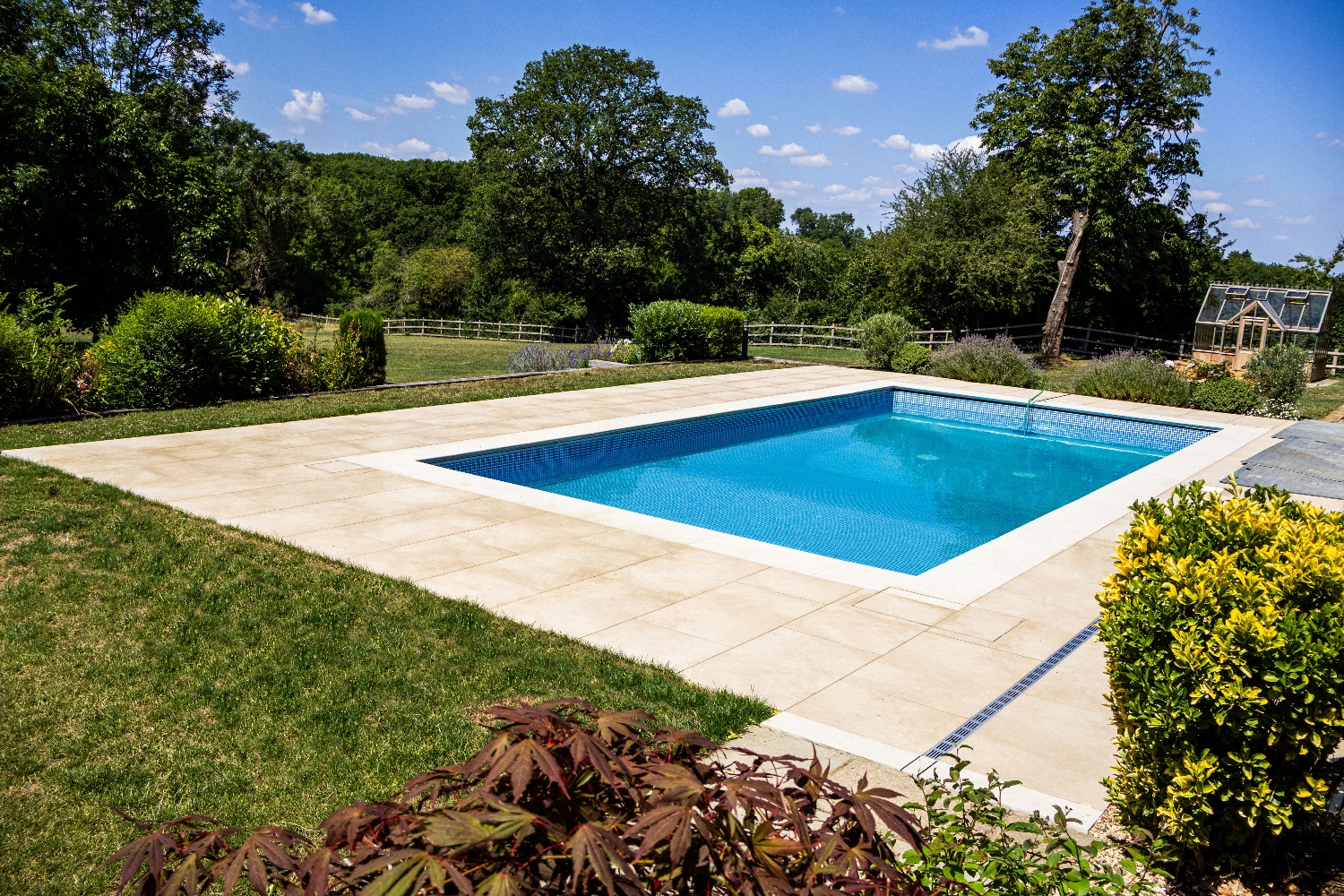 A sparkling blue swimming pool surrounded by a stone patio and vibrant greenery, under a clear blue sky with fluffy clouds.